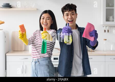 Joyeux couple asiatique avec des fournitures pour le nettoyage posant dans la cuisine Banque D'Images