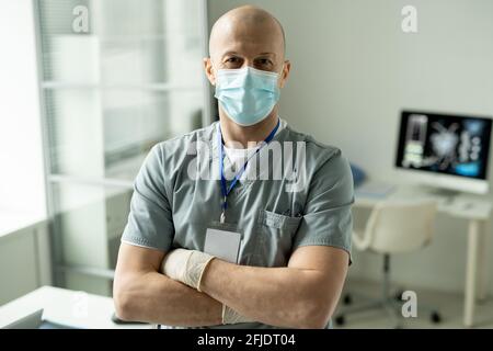 Portrait du contenu spécialiste médical expérimenté dans le masque et les exfoliants porter le badge dans le cou debout avec les bras croisés dans le bureau Banque D'Images