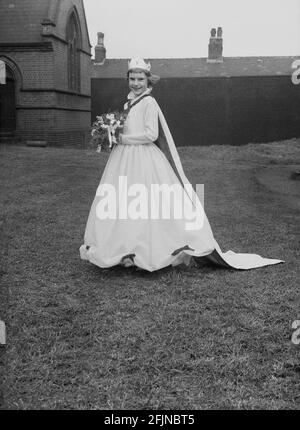 1956, historique, à l'extérieur dans le domaine de l'église, debout pour sa photo, la Reine de mai de la ville, une jeune fille portant une couronne et une longue robe, avec bouquet de fleurs. Angleterre, Royaume-Uni. Un ancien festival célébrant l'arrivée du printemps, le jour de mai a impliqué le couronnement d'une reine de mai et la danse autour d'un Maypole, des activités qui ont eu lieu en Angleterre pendant des siècles. Sélectionné parmi les filles de la région, la Reine de mai commencerait la procession des chars et de la danse. Dans le Nord industrialisé de l'Angleterre, les écoles du Dimanche de l'Église dirigeaient souvent son organisation. Banque D'Images