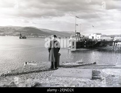 1956, historique, un homme debout à côté d'un lac dans les îles occidentales, observant les passagers à bord du bateau à vapeur, Lochfyne de Glasgow, Scottish Highlands, Écosse, Royaume-Uni. Construit par William Denny et Brothers, MV Lochfyne était un steamer MacBrayne, construit en 1931 pour le service West Highland, le premier navire de passagers côtier britannique à propulsion diesel-électrique. En 1958, elle est devenue le bateau Ardrishaig toute l'année. Elle a été retirée du service en 1969. Banque D'Images