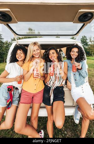Groupe de femmes diverses tenant des cocktails tout en se tenant à l'arrière de la camionnette de camping. Quatre femelles avec des bouteilles debout à une voiture pendant les vacances d'été. Banque D'Images