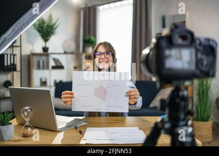 Bonne étudiante en lunettes et micro-casque, ayant une leçon en ligne tout en restant à la maison. Femme caucasienne utilisant un ordinateur portable et un appareil photo modernes pour l'apprentissage à distance. Banque D'Images