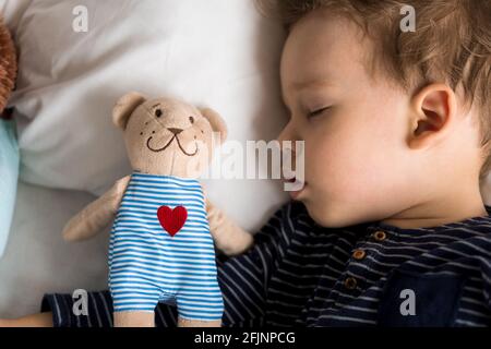 Portrait authentique malade mignon petit petit garçon d'âge préscolaire caucasien dans le sommeil bleu avec l'ours en peluche sur le lit blanc. Enfant reposant à l'heure du déjeuner. Soins Banque D'Images