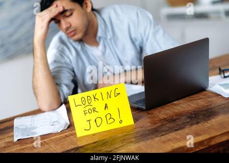 Épuisé chômeur hispanique gars, assis à la table, il ya un signe sur la table avec l'inscription à la recherche d'emploi, espérant obtenir un emploi de rêve, sentir le stress, fermer les yeux, fatigué de chercher Banque D'Images