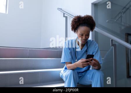 Souriant race mixte femme médecin avec masque facial à l'aide d'un smartphone assis dans les escaliers de l'hôpital Banque D'Images