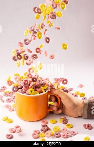 les anneaux de céréales de couleur tombent dans une tasse d'orange tenue par une main d'enfant sur fond blanc avec éclairage latéral Banque D'Images