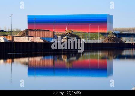 Wustermark, Allemagne. 21 avril 2021. Un véhicule de construction et un bâtiment HavelPort se reflètent dans l'eau du canal Havel. Le port intérieur, fondé en 2013, est situé au Güterverteilzentrum (GVZ) et offre de l'espace pour trois navires en même temps que son quai de 400 mètres de long. La façade de couleur arc-en-ciel dissimule le centre de distribution de la chaîne dm. Tous les magasins dm de l'est de l'Allemagne sont fournis avec des marchandises d'ici. Credit: Soeren Stache/dpa-Zentralbild/dpa/Alay Live News Banque D'Images