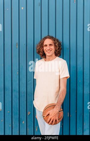 portrait vertical d'un jeune homme caucasien regardant l'appareil-photo sourire et tenant un basket-ball. Il a de longs cheveux rouges et porte des vêtements d'été décontractés. Elle est penchée sur une porte en métal bleu Banque D'Images