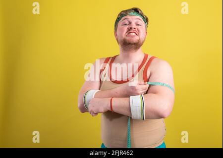 Un drôle, gai gros homme dans les vêtements de sport mesure ses bras. L'homme en surpoids entre pour le sport. Arrière-plan jaune isolé Banque D'Images