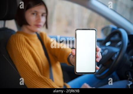 Une jeune femme heureuse conduisant une voiture, elle montre un écran de téléphone vide . Applications mobiles utiles pour le conducteur. Vue latérale à l'intérieur de la voiture, maquette Banque D'Images