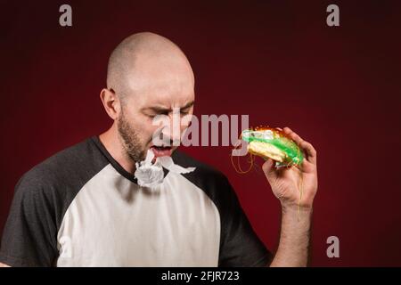 Le concept de restauration rapide et d'écologie. Un homme chauve avec une barbe, tenant un hamburger et cracher plastique de remplissage. Arrière-plan Bourgogne. Banque D'Images