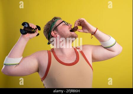 drôle d'homme gras dans des vêtements de sport avec des haltères dans son les mains mangent des biscuits au chocolat Banque D'Images