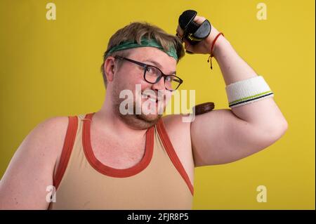 drôle d'homme gras dans des vêtements de sport avec des haltères dans son les mains mangent des biscuits au chocolat Banque D'Images