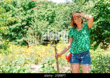 Une jeune femme caucasienne avec une fourche dans ses mains pose sur un fond de végétation estivale luxuriante et d'arbres. Jour d'été ensoleillé. Concept de gar Banque D'Images