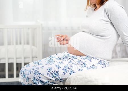 Femme enceinte avec une cigarette dans les mains, assise sur le lit. Mains avec une cigarette et l'estomac gros plan. Le concept de dépendance à la nicotine et Banque D'Images