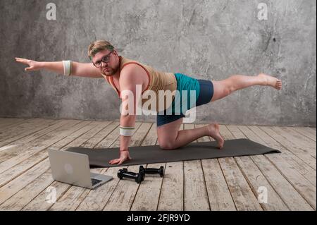 drôle d'homme gras dans les vêtements de sport faisant des exercices. Il joue du sport à distance tout en regardant son ordinateur portable Banque D'Images