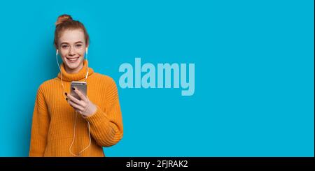Ginger dame avec des taches de rousseur sourit à l'appareil photo sur un mur de studio bleu pour écouter de la musique sur des écouteurs Banque D'Images