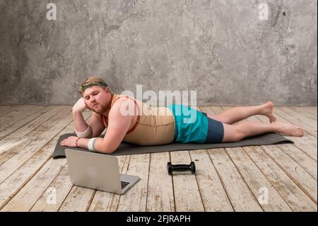 drôle d'homme gras faisant du sport sur le tapis de yoga, faisant du sport en ligne, regardant dans l'ordinateur portable Banque D'Images