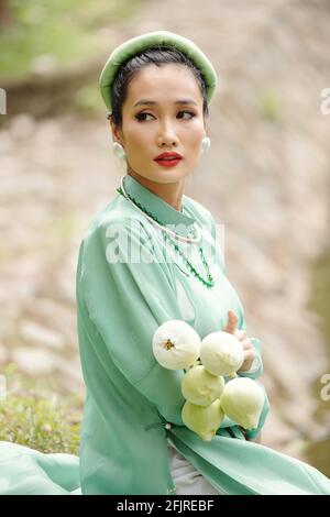 Portrait de la jeune femme sérieuse en robe de menthe légère et coiffures de repos à l'extérieur avec bouquet de fleurs de lotus dans les mains Banque D'Images