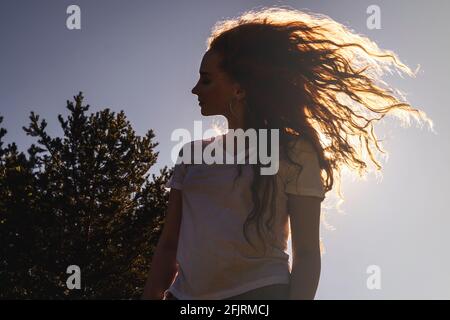 Silhouette sombre de jeune femme heureuse avec de longs cheveux bouclés au soleil. Fond ciel. Banque D'Images