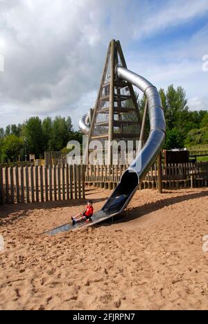 Grande structure à quatre côtés avec jeune garçon en rouge émergeant d'un tube fermé incurvé agissant comme toboggan pour enfants, Telford Town Park, Shropshire, Angleterre Banque D'Images