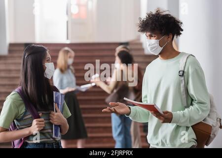 Étudiants multiethniques dans des masques médicaux tenant des carnets et parlant Banque D'Images