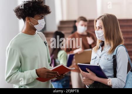 Étudiants multiethniques dans des masques médicaux tenant des carnets dans l'université Banque D'Images