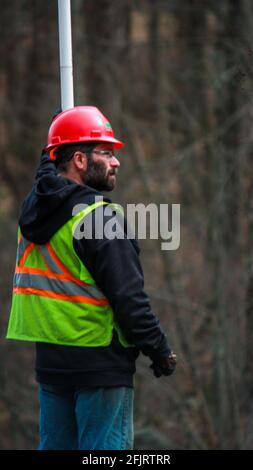 MIDDLETOWN, NY, ÉTATS-UNIS - 15 mars 2019: Travailleur de la construction en Red HardHat tient le poteau dirigeant la circulation portant jaune gilet réfléchissant sur un bois Banque D'Images