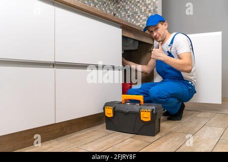 Le handyman en combinaison répare les tuyaux sous l'évier dans la cuisine, à côté d'une boîte d'outils. Regarde l'appareil photo et montre l'affiche du pouce vers le haut Banque D'Images