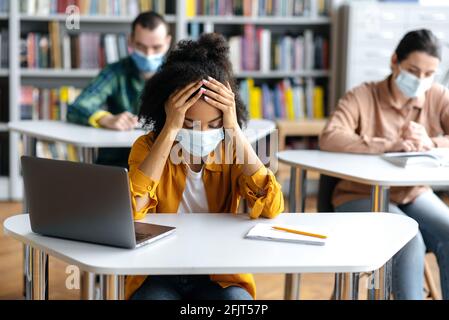 Éducation pendant une pandémie. Frustrée et surtravaillée, la jeune fille étudiante afro-américaine portant un masque médical protecteur est assise à une table de la bibliothèque, se préparant aux examens, fatiguée, les camarades de classe sont assis à l'arrière-plan Banque D'Images