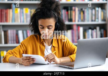 Étudiante afro-américaine concentrée avec micro-casque, dans des vêtements élégants, assise à table dans la bibliothèque de l'université, à l'aide d'un ordinateur portable, regardant l'ordinateur portable avec concentration, prenant des notes, préparant un examen Banque D'Images