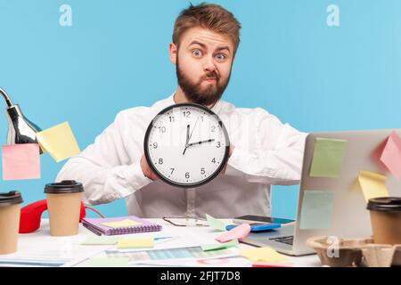 Drôle d'homme malheureux avec une barbe en chemise blanche montrant une grande horloge murale assis sur le lieu de travail, tous couverts de notes collantes, pressé de terminer son travail. Po Banque D'Images