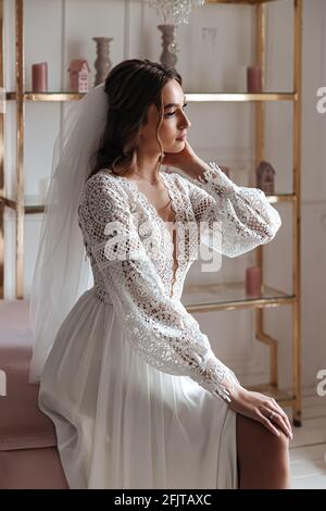 Mariée dans une robe de mariage dans le style boho et avec un voile posant assis dans une pièce confortable. Séance photo de mariage, matin de la mariée. Banque D'Images