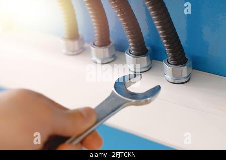 Il s'agit de câbles d'alimentation en isolant ondulé et d'un électricien de roche avec une clé à proximité d'un mur bleu. Connexion fiable et correcte des fils au chemin de câbles. Photo publicitaire pour le service d'alimentation. Banque D'Images