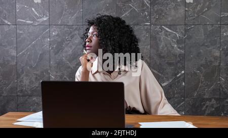 Une femme afro-américaine, responsable de l'entreprise, aux cheveux bouclés, est assise à table avec ordinateur portable et des papiers donnant sur la fenêtre dans le bureau avec mur en marbre Banque D'Images