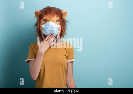 Jeune femme portant un lion et un masque médical de protection, couvrant la bouche avec la main, isolée sur fond bleu. Banque D'Images