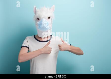 Jeune femme portant l'alpaga et un masque de protection médicale, garde le pouce vers le haut geste d'approbation, isolé sur fond bleu. Banque D'Images