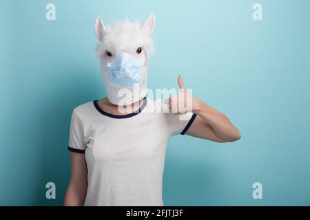 Jeune femme portant l'alpaga et un masque de protection médicale, garde la main raillée pouce vers le haut geste d'approbation, isolé sur fond bleu. Banque D'Images