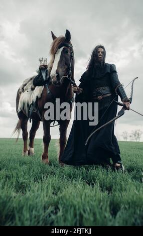 Femme en chaîne de courrier à l'image de la guerrière médiévale se tient avec l'arc dans sa main près du cheval dans le champ. Banque D'Images