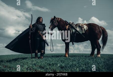 Femme en chaîne de courrier à l'image de la guerrière médiévale se tient avec l'arc dans sa main près du cheval dans le champ. Banque D'Images