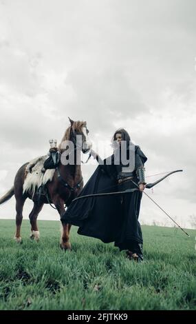 Femme en chaîne de courrier à l'image de la guerrière médiévale se tient avec l'arc dans sa main près du cheval dans le champ. Banque D'Images