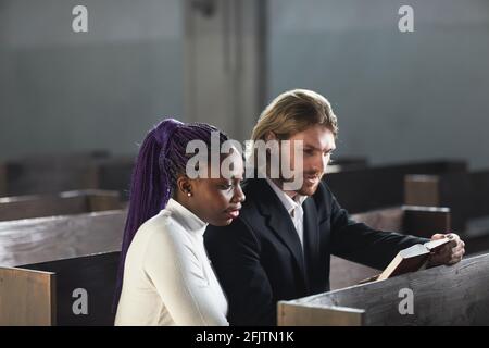 Jeune couple multiethnique assis sur le banc ensemble et lisant La Bible ensemble dans l'église Banque D'Images