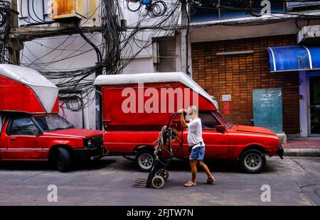 Un vieil homme thaïlandais pousse un chariot vide le long de la rue tout en passant deux vieux véhicules Mazda garés dans le quartier chinois de Bangkok, en Thaïlande Banque D'Images