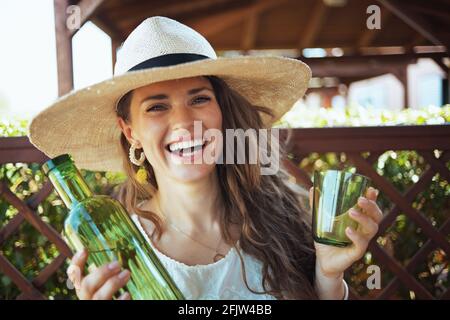 Portrait de la jeune femme heureuse en chemise blanche avec bouteille d'eau, verre et chapeau dans le patio. Banque D'Images