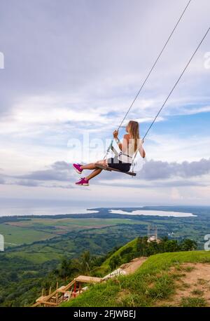 Une jeune fille, blonde, se balançant sur une balançoire sur une pente de montagne en été. Balancer haut dans les montagnes au-dessus de la vallée. République dominicaine. Réglage du soleil Banque D'Images