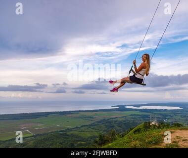 Une jeune fille, blonde, se balançant sur une balançoire sur une pente de montagne en été. Balancer haut dans les montagnes au-dessus de la vallée. République dominicaine. Réglage du soleil Banque D'Images