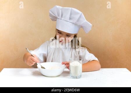 Une petite fille habillée comme un cuisinier pétrit la pâte. Concept de mode de vie enfant de cuisine. Le gamin aime, s'amuse, étudie et joue dans la cuisine Banque D'Images