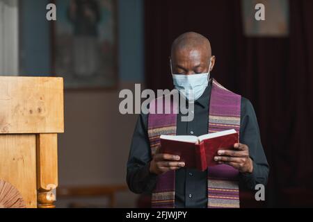 Prêtre africain en masque et en costume lisant la Bible pendant la messe dans l'église Banque D'Images