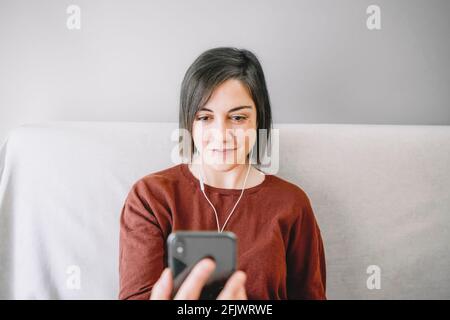 Portrait d'une femme ayant un appel vidéo avec son smartphone noir et des écouteurs blancs et assis sur le canapé près du fenêtre lumineuse dans son salon à Banque D'Images