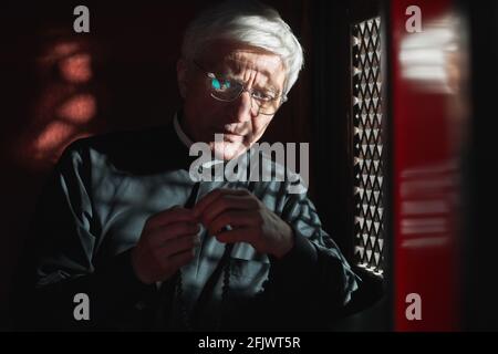 Prêtre senior assis avec des perles rosaires assis dans confessionnel et écouter la confession Banque D'Images
